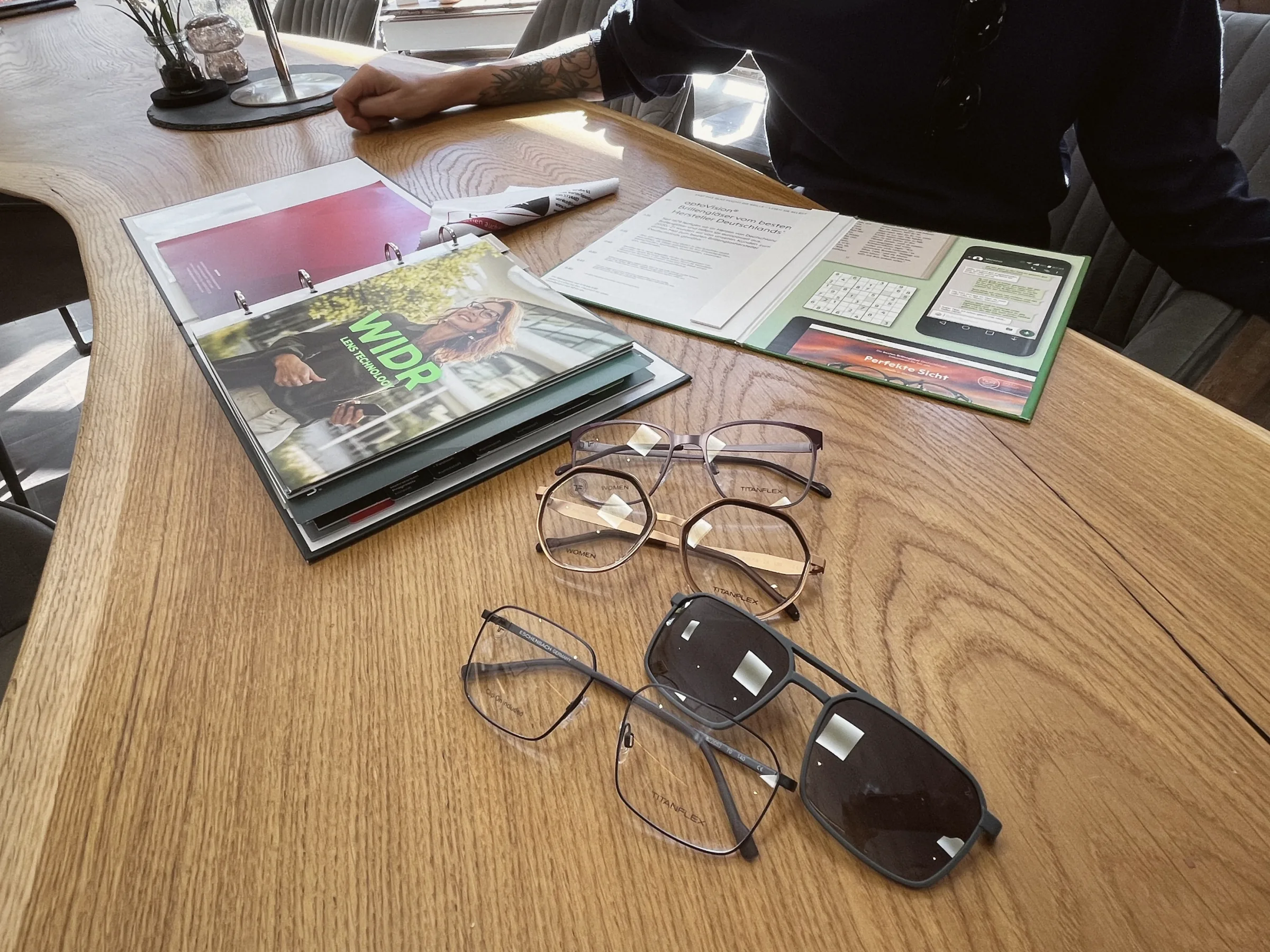 Mehrere Brillenfassungen und eine Sonnenbrille liegen bei der Beratung auf einem Holztisch neben Brillenglas-Katalogen.