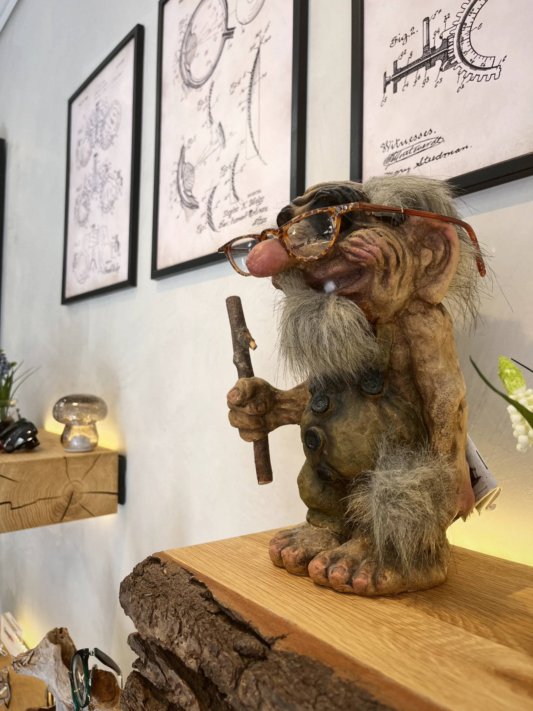 Eine Troll-Figur mit Brille und Wanderstock steht im Laden vor gerahmten Zeichnungen.